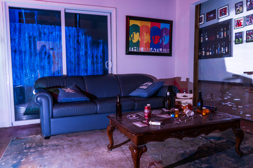 Living room within Decision Tactical's indoor house scenario in North Orlando, featuring a leather sofa, a sliding glass door, and a coffee table cluttered with beer bottles, pill bottles, and drug paraphernalia props set up for law enforcement scenario training.